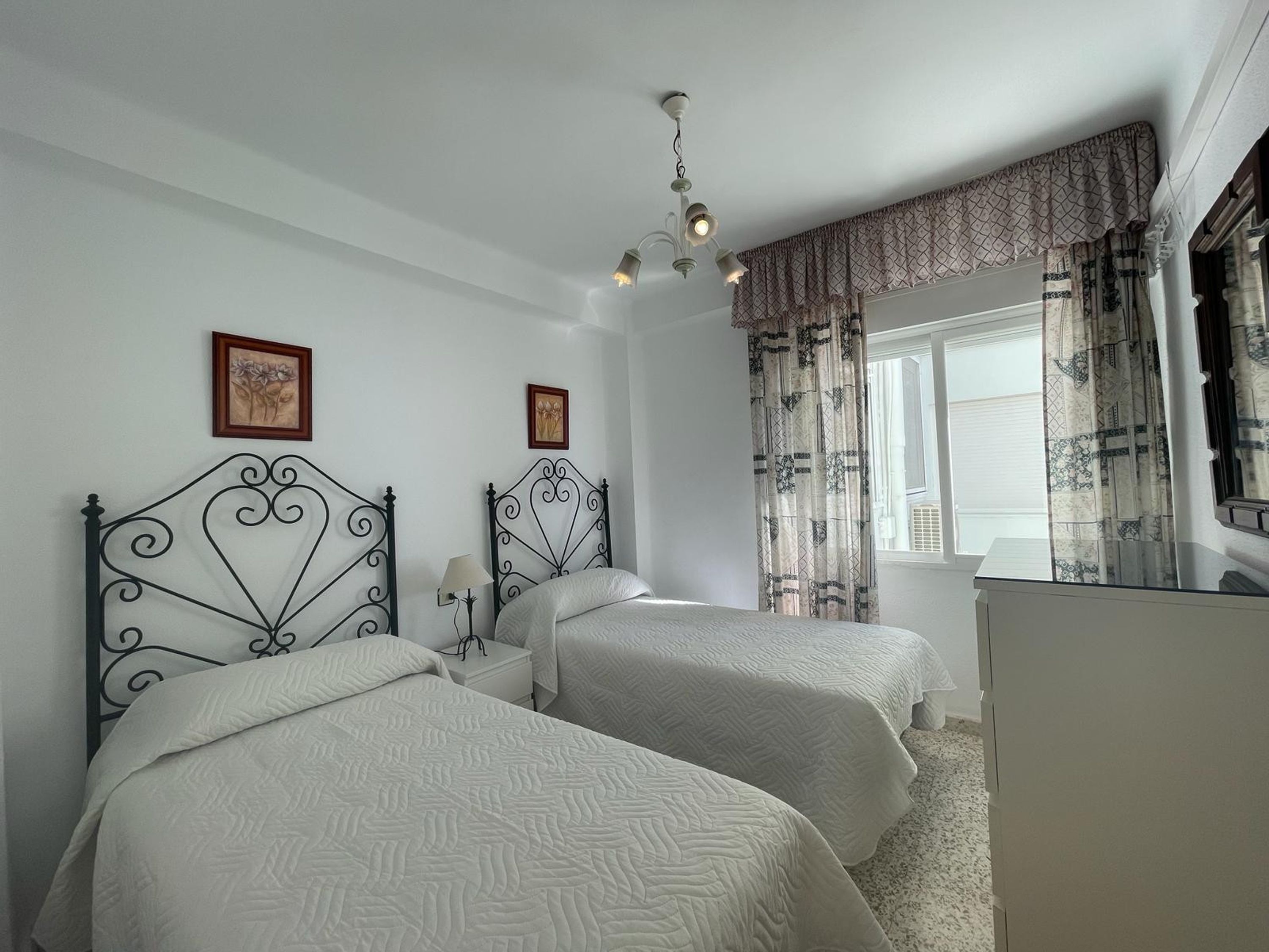 A bedroom with two beds, a ceiling fan, a window with patterned curtains, and a dresser.