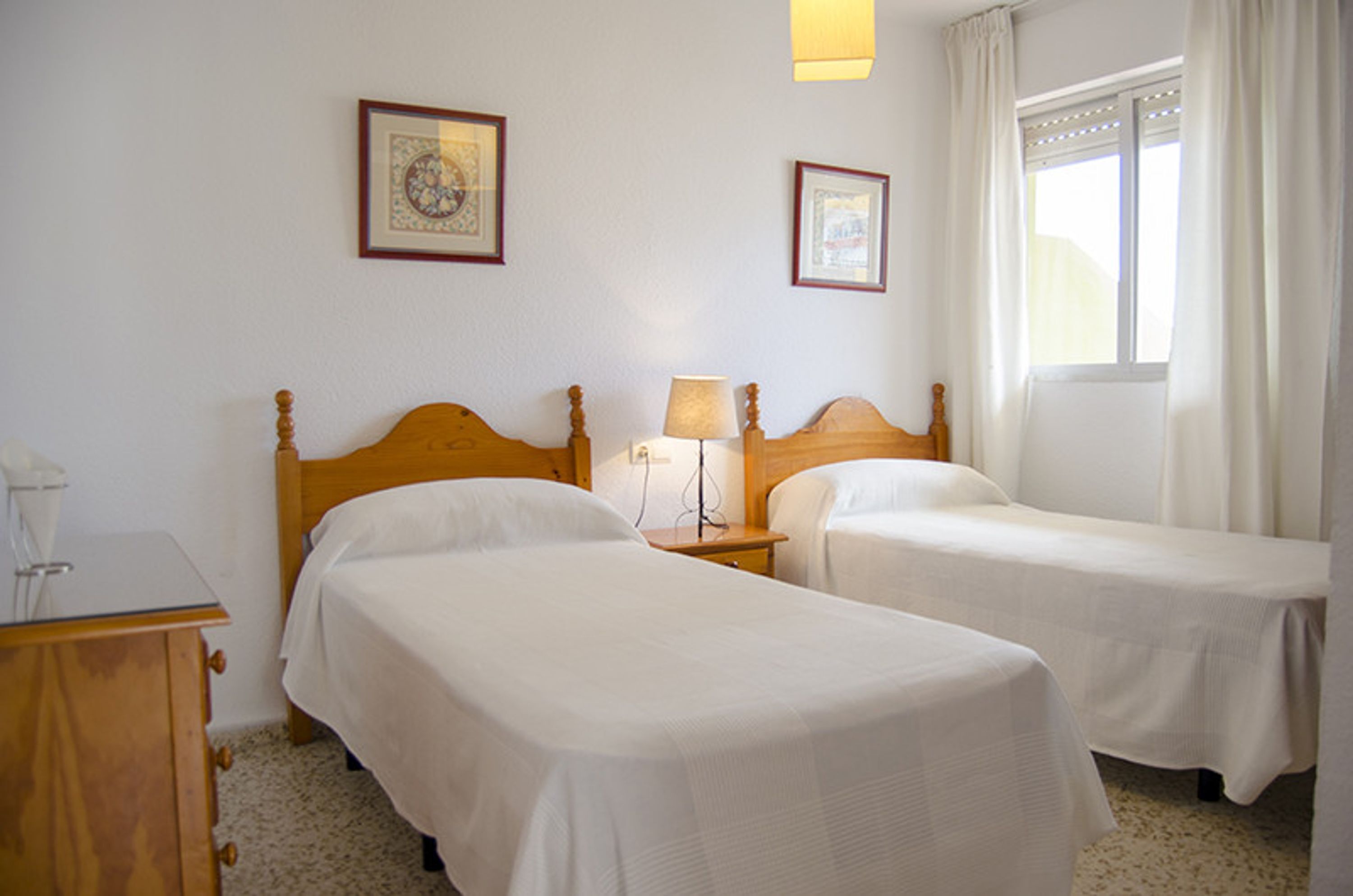 A hotel room with two single beds, a bedside table with a lamp, a wooden dresser, and framed pictures on the wall.