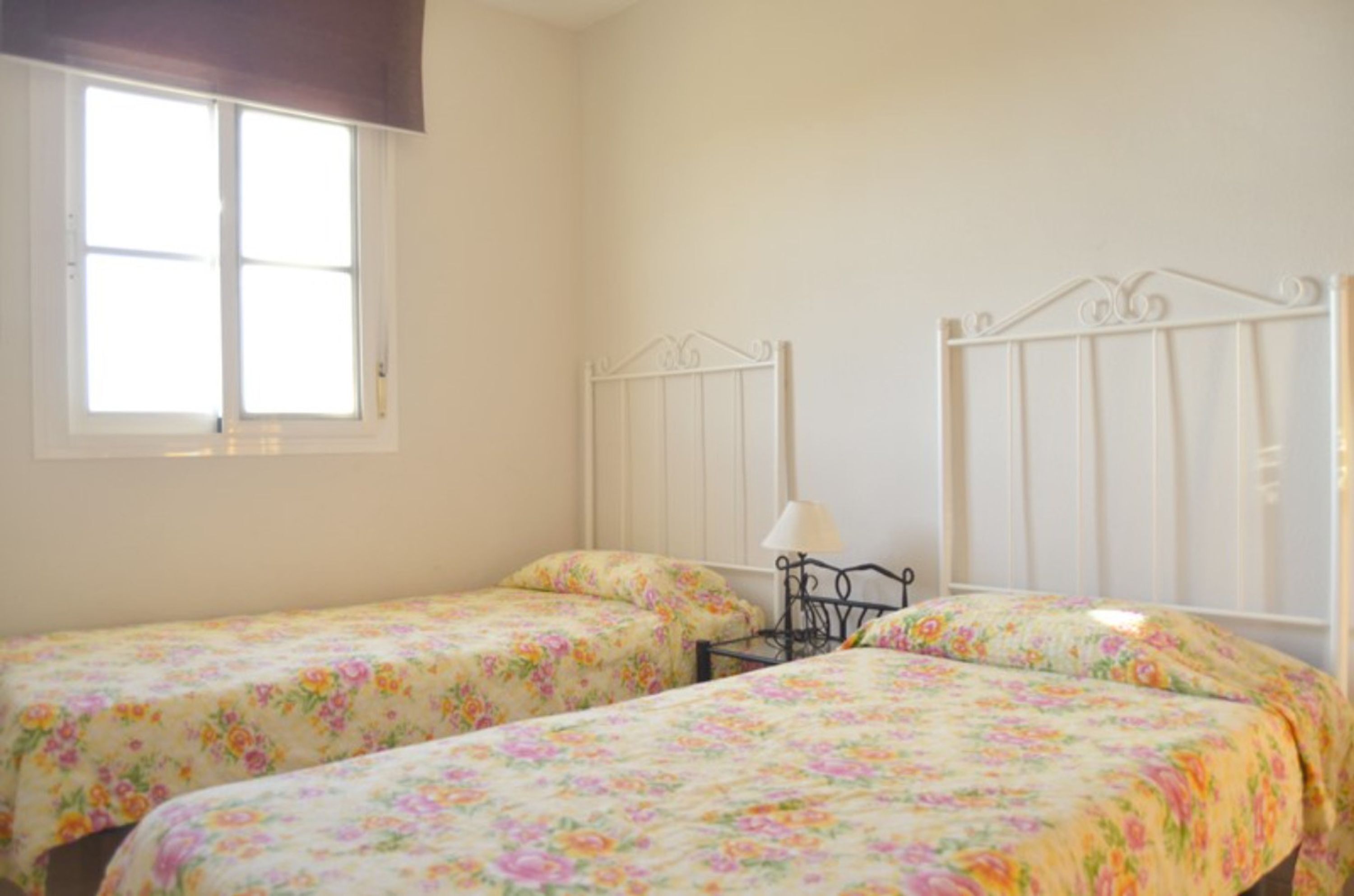Een kamer met twee enkele bedden, bloemenbeddengoed, een raam en een wit metalen hoofdeinde.