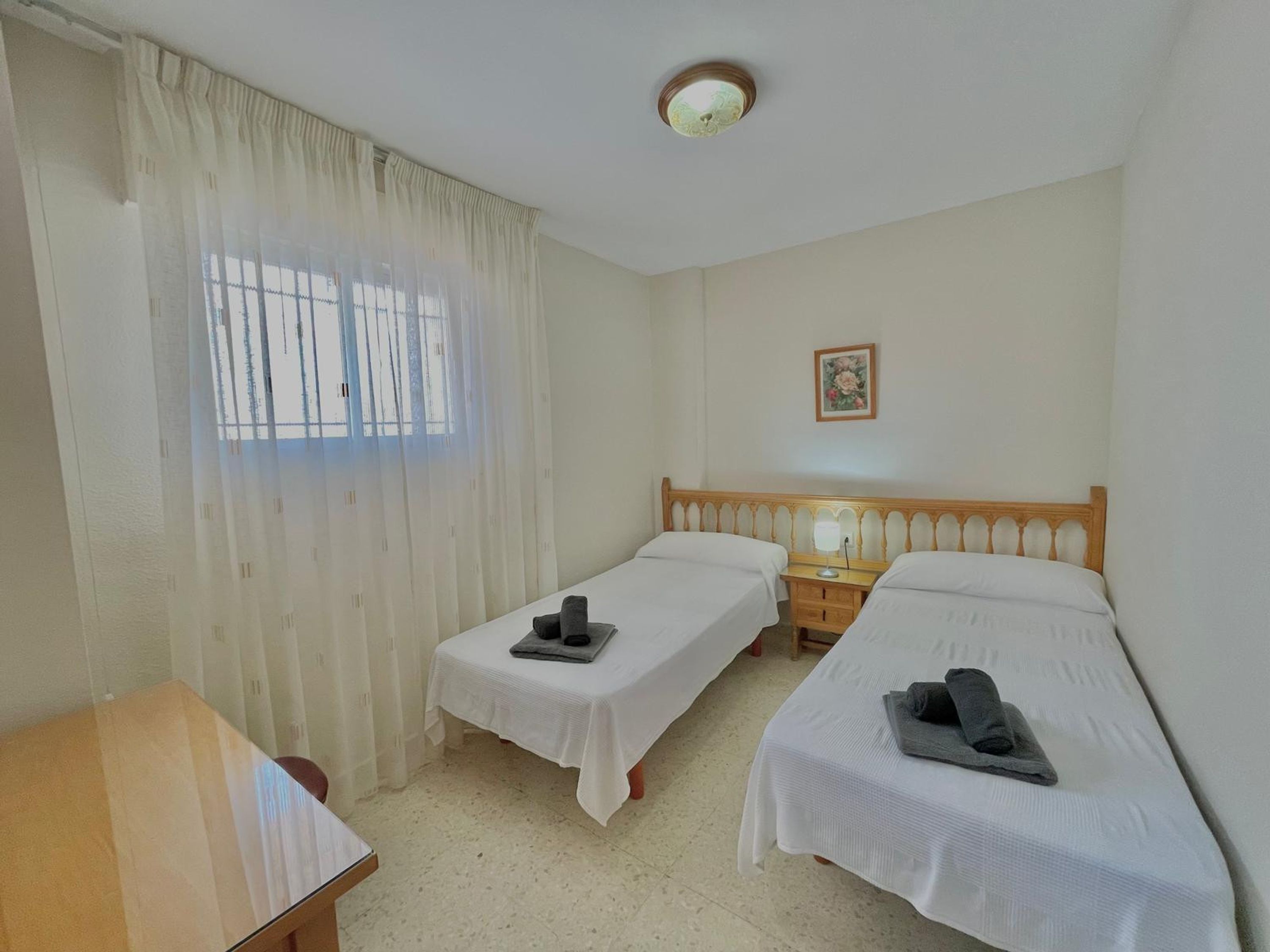 Quarto com duas camas de solteiro, uma mesa de madeira, uma pequena escrivaninha, um criado-mudo, um abajur e uma janela com cortinas leves.