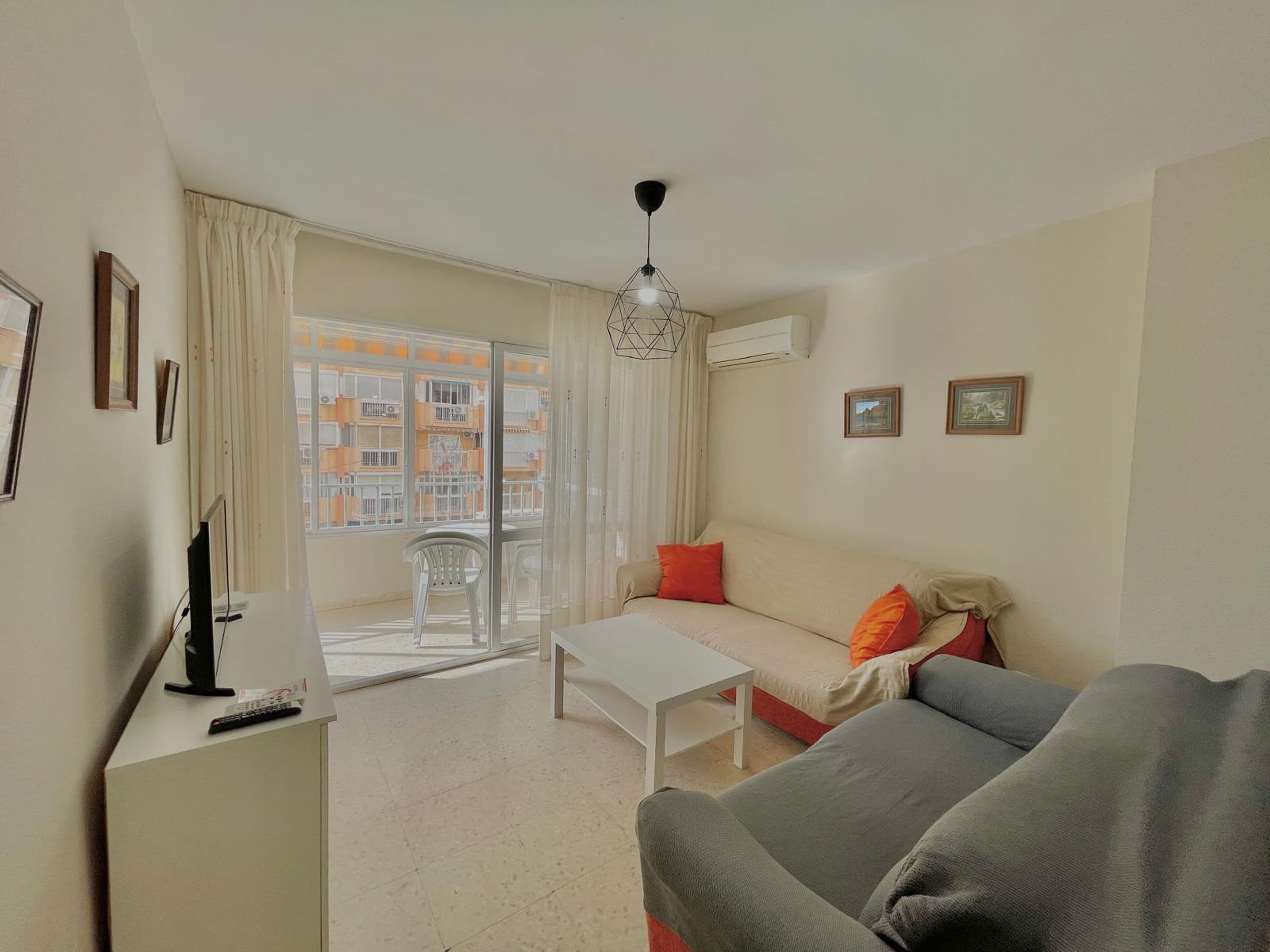 Una sala de estar con sofá, mesa de centro, televisor y balcón con vistas a edificios.