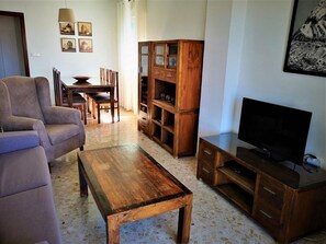A living room with a sofa, coffee table, wooden cabinet, TV stand, and a dining area with chairs and a table. - Terrasol Atico Azucarera 5I (Velez-Malaga)