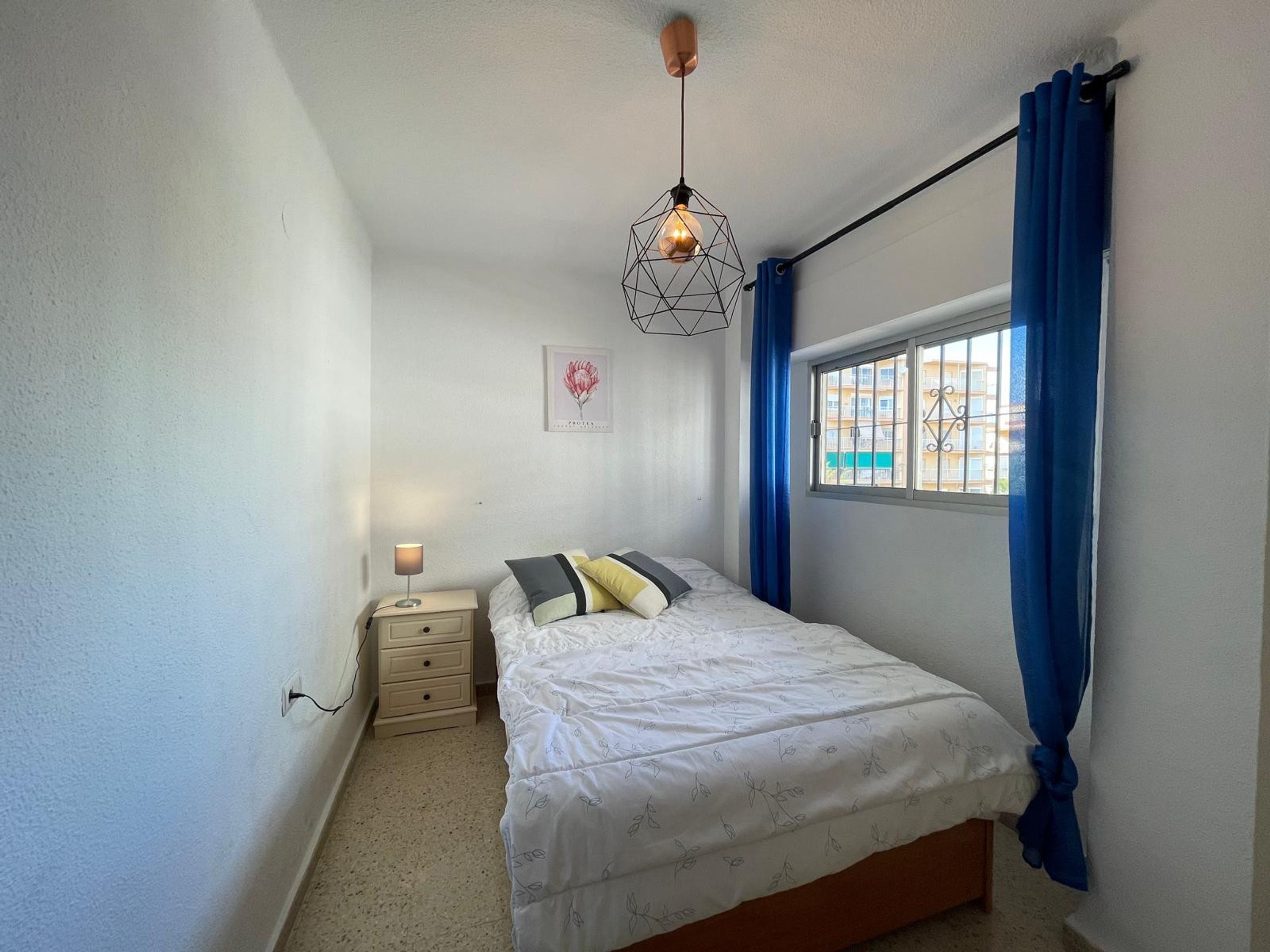 A bedroom with a bed, a nightstand, a wall-mounted lamp, a window with blue curtains, and a cityscape view.