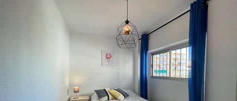 A bedroom with a bed, a nightstand, a wall-mounted lamp, a window with blue curtains, and a cityscape view.