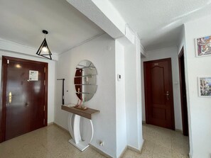 A hallway with two wooden doors, a white wall with a round mirror, a small white console table, and a ceiling light fixture. - Terrasol Arenas 4 in Torre del Mar (Velez-Malaga)