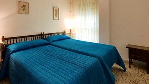Un dormitorio con dos camas, una mesita de noche, una lámpara, una ventana con cortinas y un cuadro colgado en la pared.