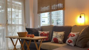 Un salon avec un canapé, une petite table en bois sur laquelle sont disposés des fruits, un tapis tissé, une fenêtre avec des stores et un appareil de climatisation.
