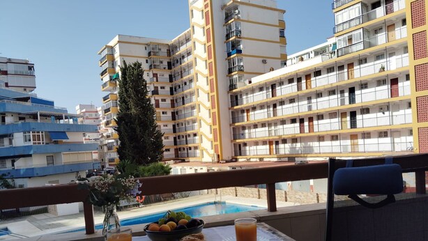 A balcony with a table set for breakfast, a pool, and high-rise buildings in the background. - Terrasol Estrella 2 in Torre del Mar (Velez-Malaga)