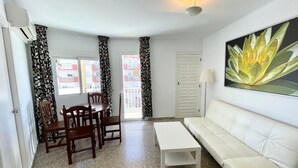 A living room with a white sofa, a dining table with chairs, a white coffee table, patterned curtains, and a painting of a flower. - Terrasol Trinidad 4 in Torre del Mar (Velez-Malaga)