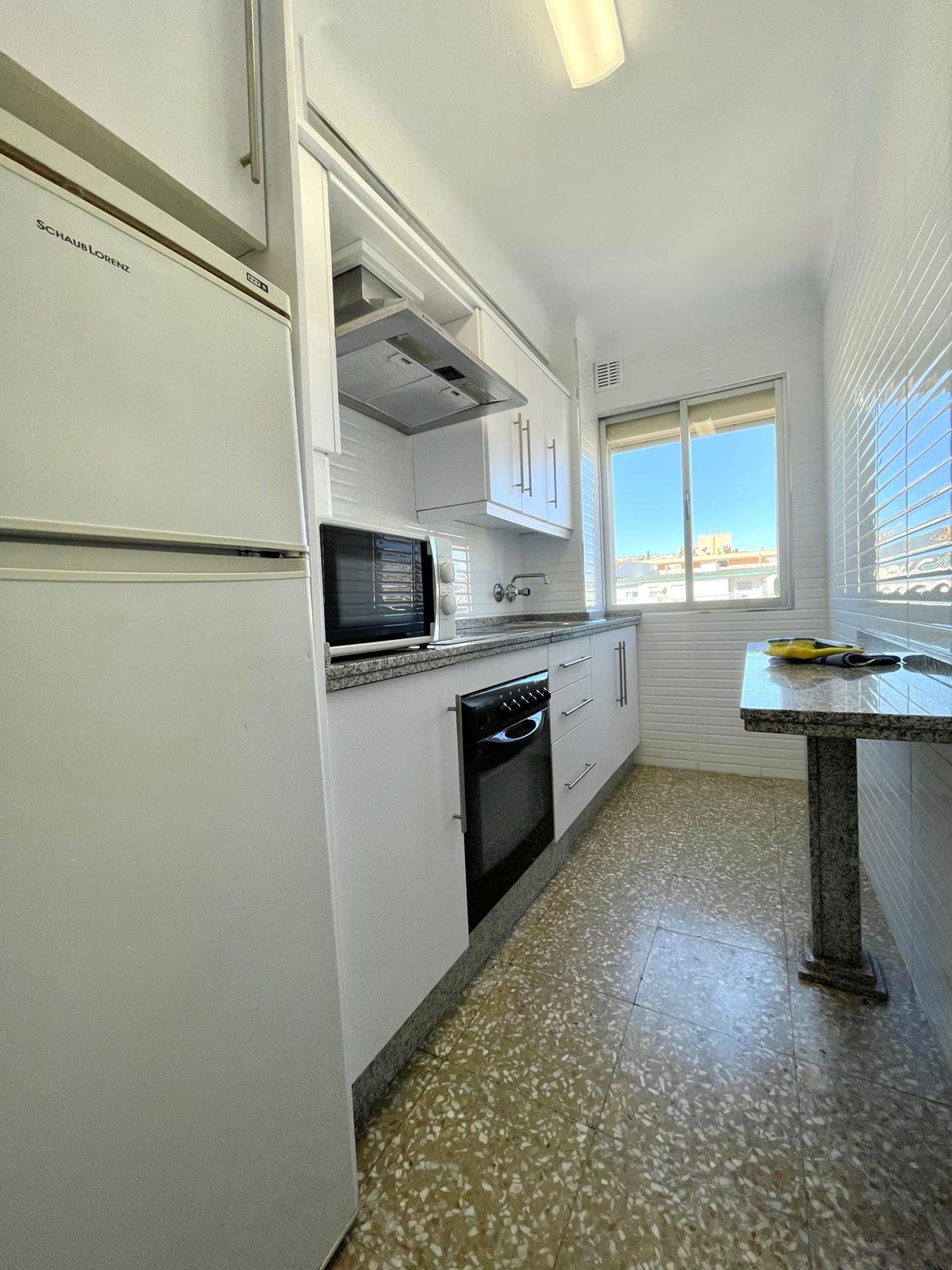 Cozinha compacta com armários brancos, micro-ondas embutido, fogão e janela com vista para a paisagem urbana.