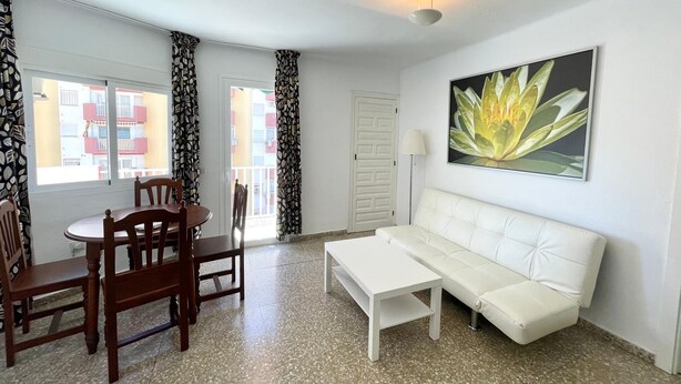 A room with a dining table, chairs, a white sofa, a floor lamp, and a large window with patterned curtains. - Terrasol Trinidad 4 in Torre del Mar (Velez-Malaga)
