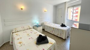 A bedroom with two beds, a nightstand with a lamp, a wardrobe, and a window with a view of buildings. - Terrasol Trinidad 4 in Torre del Mar (Velez-Malaga)