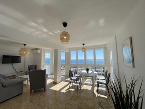 Amplia sala de estar con un gran ventanal con vista al mar, una mesa de comedor con sillas, un sofá gris y un televisor de pantalla plana montado en la pared.