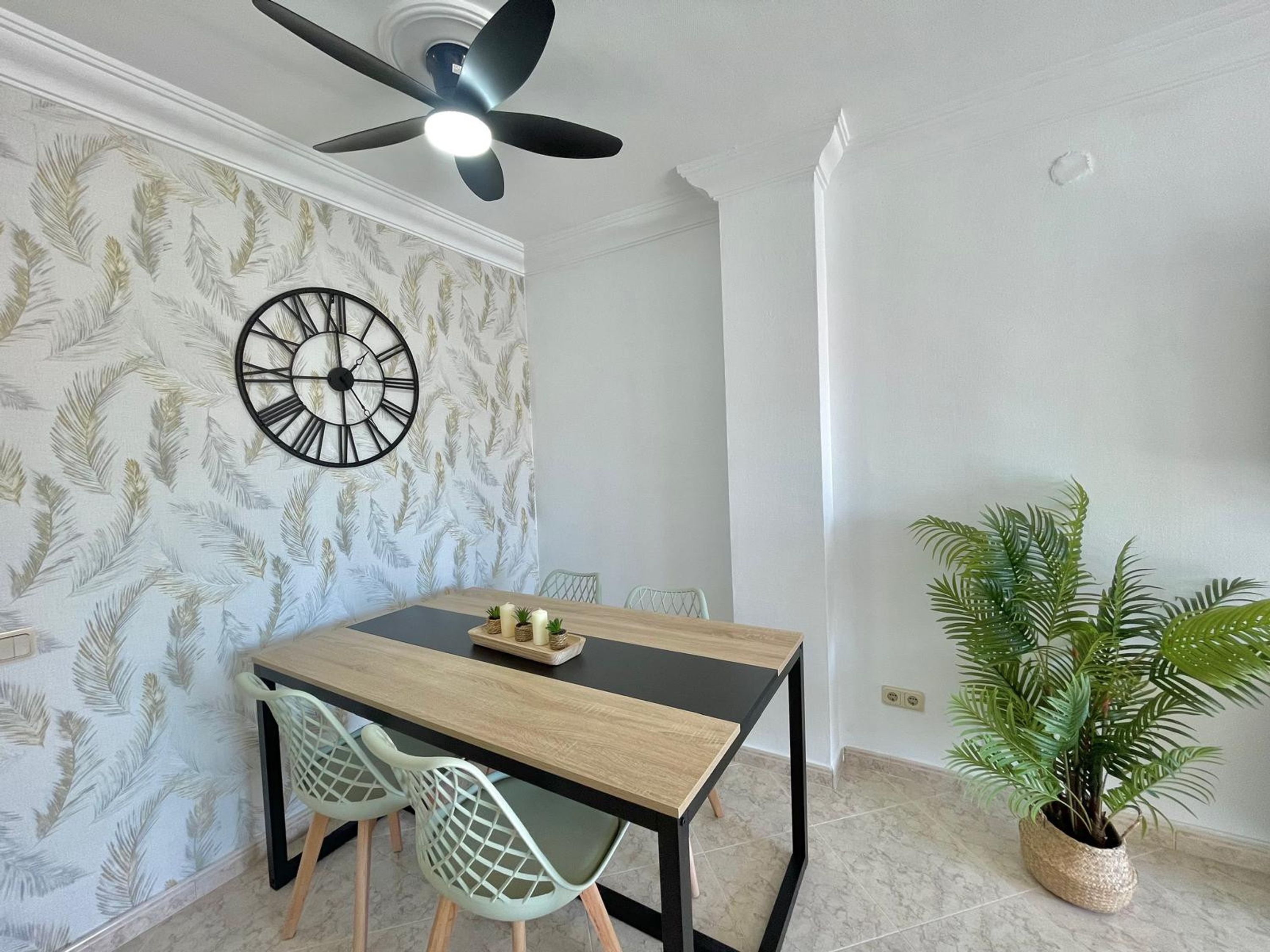 Un comedor con una mesa de madera, dos sillas, un reloj de pared, un ventilador de techo y una planta en maceta.