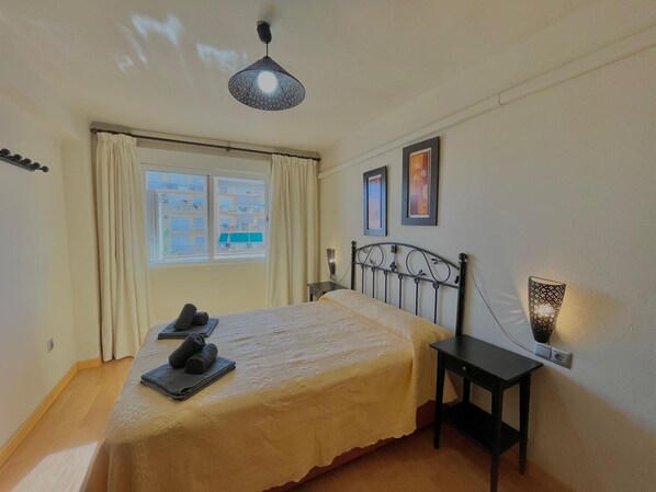 Une chambre à coucher comprenant un lit, une table de chevet, une lampe, une fenêtre avec des rideaux et un luminaire au plafond.