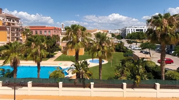A pool area with palm trees, a clear blue sky, and buildings in the background. - Terrasol Azucarera 3 Dormitorios (Velez-Malaga)