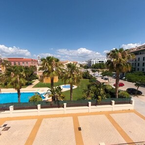 A pool area with palm trees, a clear sky, and buildings in the background. - Terrasol Azucarera 3 Dormitorios (Velez-Malaga)
