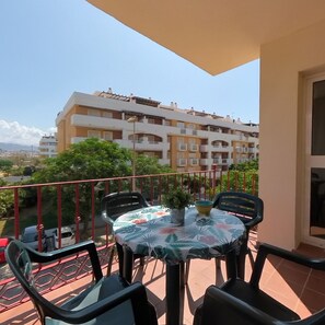 A balcony with a table and chairs, a potted plant, and a view of a multi-story building. - Terrasol Azucarera 3 Dormitorios (Velez-Malaga)
