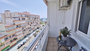 A balcony with a potted plant, a table with a bottle and glass, and a view of a multi-story building with balconies and parked cars. - Terrasol Franlou Vistas AL MAR (Velez-Malaga)