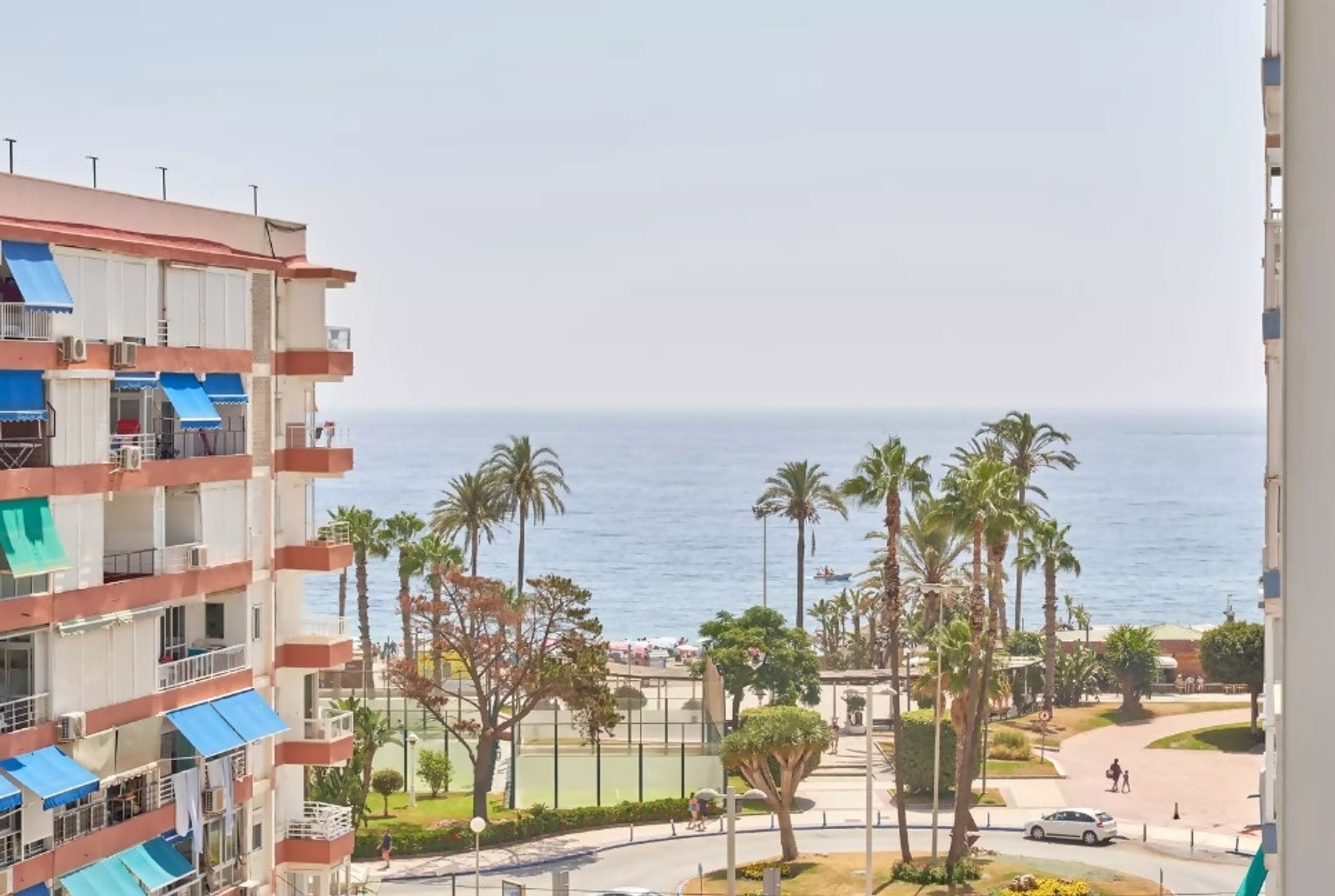 A multi-story building with balconies, a view of the sea, palm trees, and a pedestrian pathway.