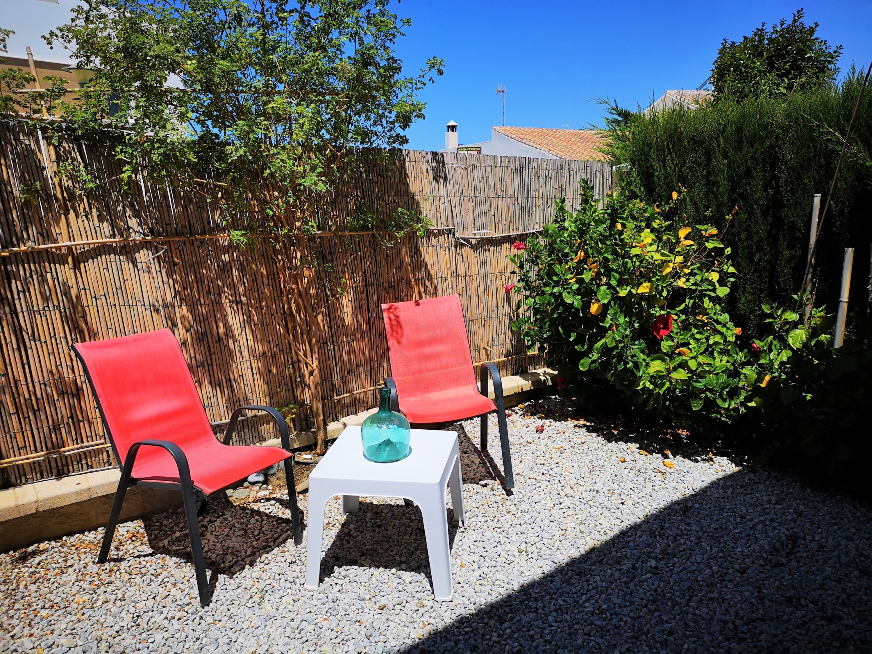 Two red chairs and a small white table in a garden area with gravel ground, bamboo fence, and a bush with red flowers.