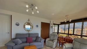 Un soggiorno con divano, poltrone, un tavolino in legno, una ventola a soffitto, uno specchio e vista sugli edifici attraverso ampie finestre.