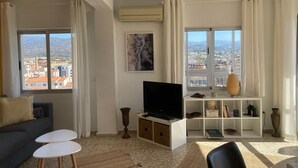 A living room with a TV, a sofa, two small tables, a bookshelf, and a view of the city through large windows. - Terrasol Roma 9 in Torre del Mar (Velez-Malaga)