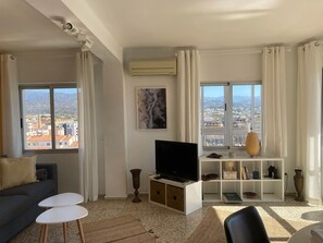A living room with a TV, a sofa, two small tables, a bookshelf, and a view of the city through large windows. - Terrasol Roma 9 in Torre del Mar (Velez-Malaga)