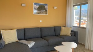A living room with a sofa, two side tables, a painting, and a window with a view. - Terrasol Roma 9 in Torre del Mar (Velez-Malaga)