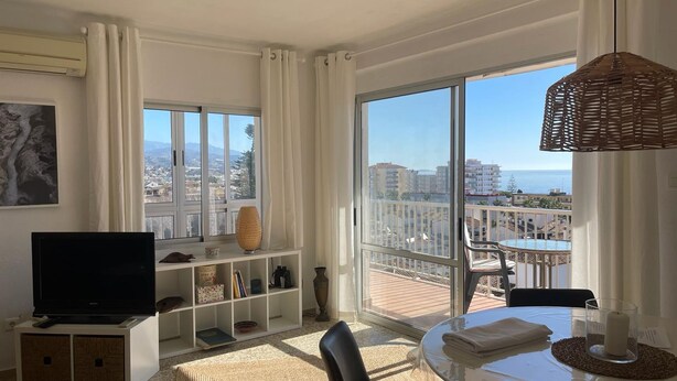 A modern living room with a large TV, a bookshelf, a dining area with a table and chairs, and a balcony with a view of the city and sea. - Terrasol Roma 9 in Torre del Mar (Velez-Malaga)