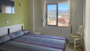 A bedroom with a bed, a chair, a window with curtains, a small table, and a mirror on the wall. - Terrasol Roma 9 in Torre del Mar (Velez-Malaga)