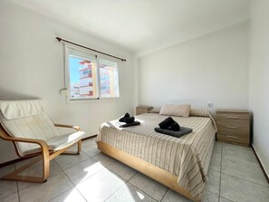 A bedroom with a bed, a chair, a nightstand, and a window with a view of buildings. - Terrasol Ahumada 1 in Torre del Mar (Velez-Malaga)