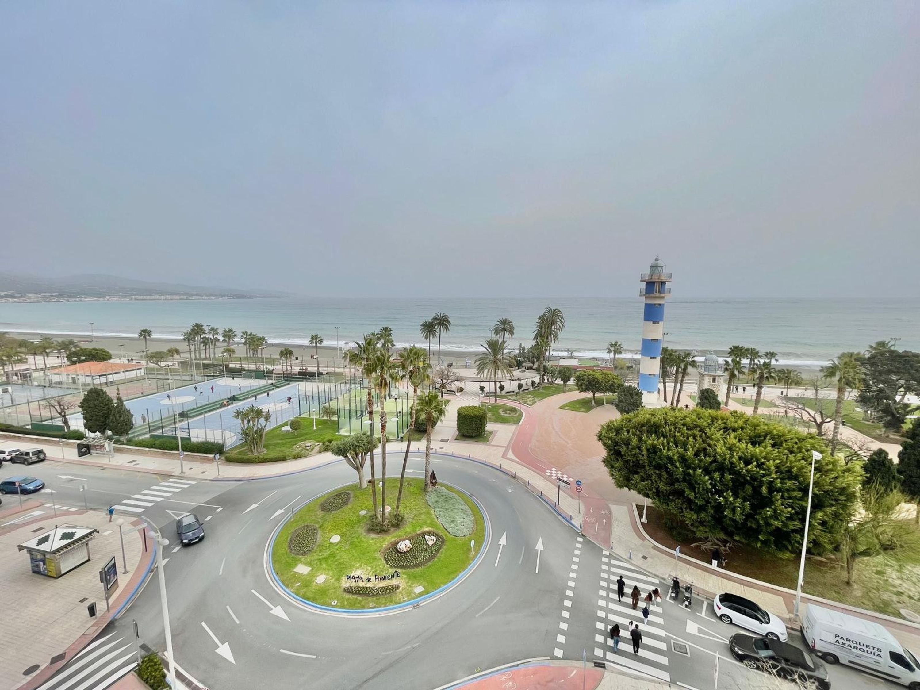 Uma rotatória com área verde central, um farol e uma praia ao fundo.
