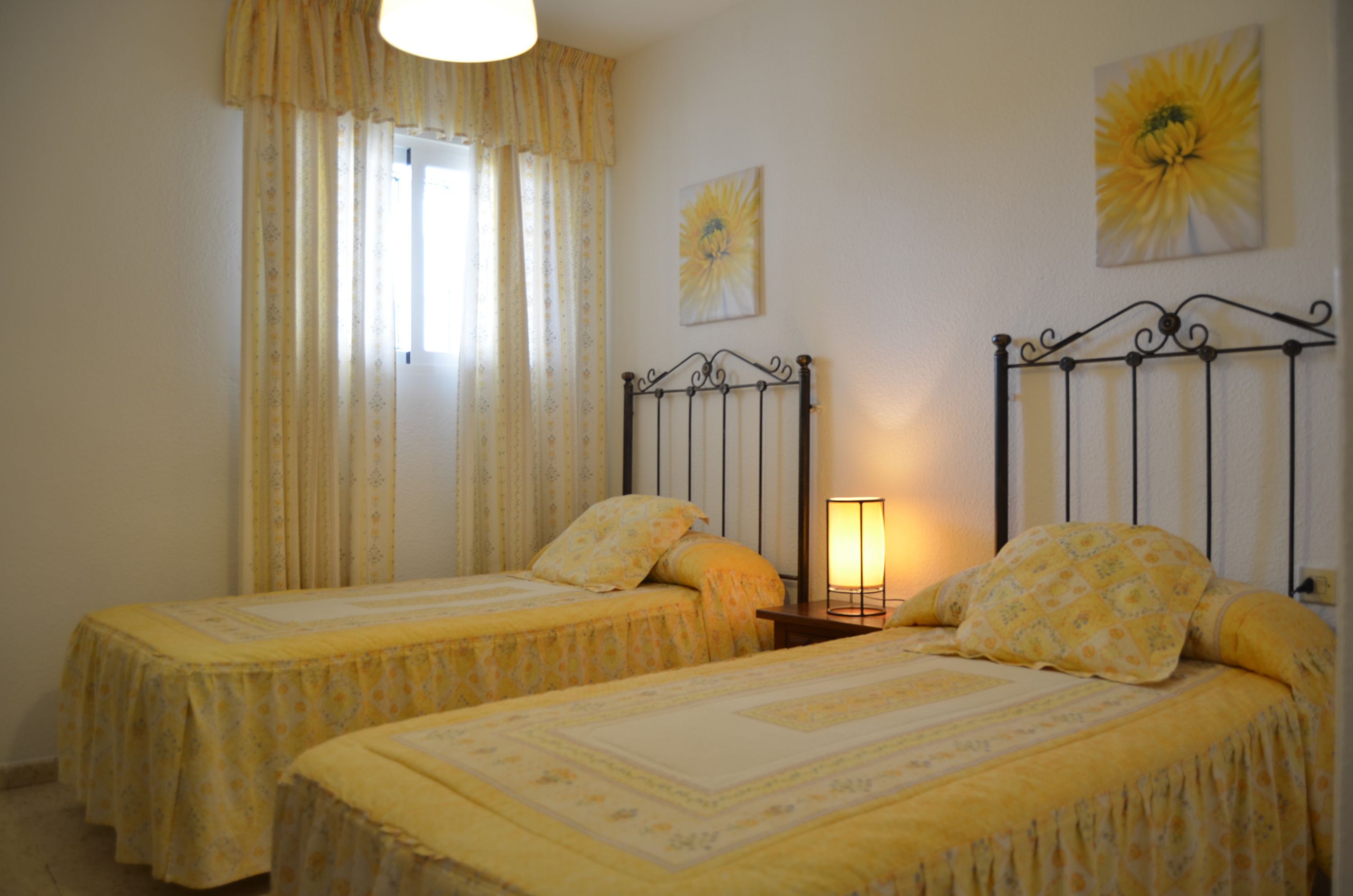 Una habitación con dos camas, una ventana con cortinas, una lámpara de pared y dos cuadros de flores.