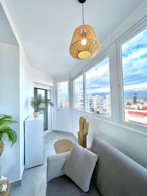 Ein modernes Wohnzimmer mit einer Couch, einem gewebten Sessel, einer kleinen Kommode und einem großen Fenster mit Blick auf die Stadt.