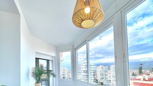 A modern living room with a sofa, a woven chair, a small cabinet, and a large window offering a city view. - Terrasol Plazamar 2-9-14 (Velez-Malaga)