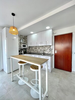 A modern kitchen with white cabinets, a wooden table, two bar stools, a hanging light fixture, and a microwave. - Terrasol Plazamar 2-9-14 (Velez-Malaga)