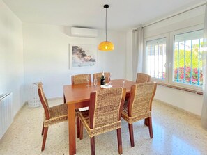 Un coin repas avec une table en bois et des chaises en osier, un luminaire suspendu, un tableau au mur et une fenêtre donnant sur un paysage verdoyant.