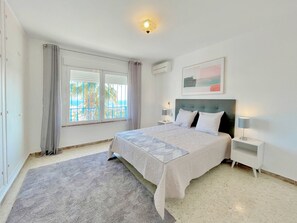 Une chambre à coucher avec un lit, deux tables de chevet, une fenêtre avec des rideaux, un appareil de climatisation et un tableau au mur.