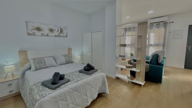 A modern bedroom with a bed, bedside lamps, a wardrobe, and a seating area with a sofa and shelves. - Terrasol Maenoba Playa 2 (Velez-Malaga)