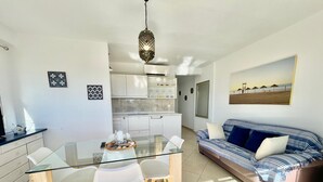 Une cuisine moderne avec des meubles blancs, un coin repas doté d’une table et de chaises en verre, et un salon avec un canapé et un tableau représentant une plage.