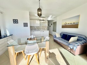 Eine moderne Küche mit weißen Schränken, ein Essbereich mit Glastisch und Stühlen sowie ein Wohnzimmer mit Sofa und einem Strandbild.