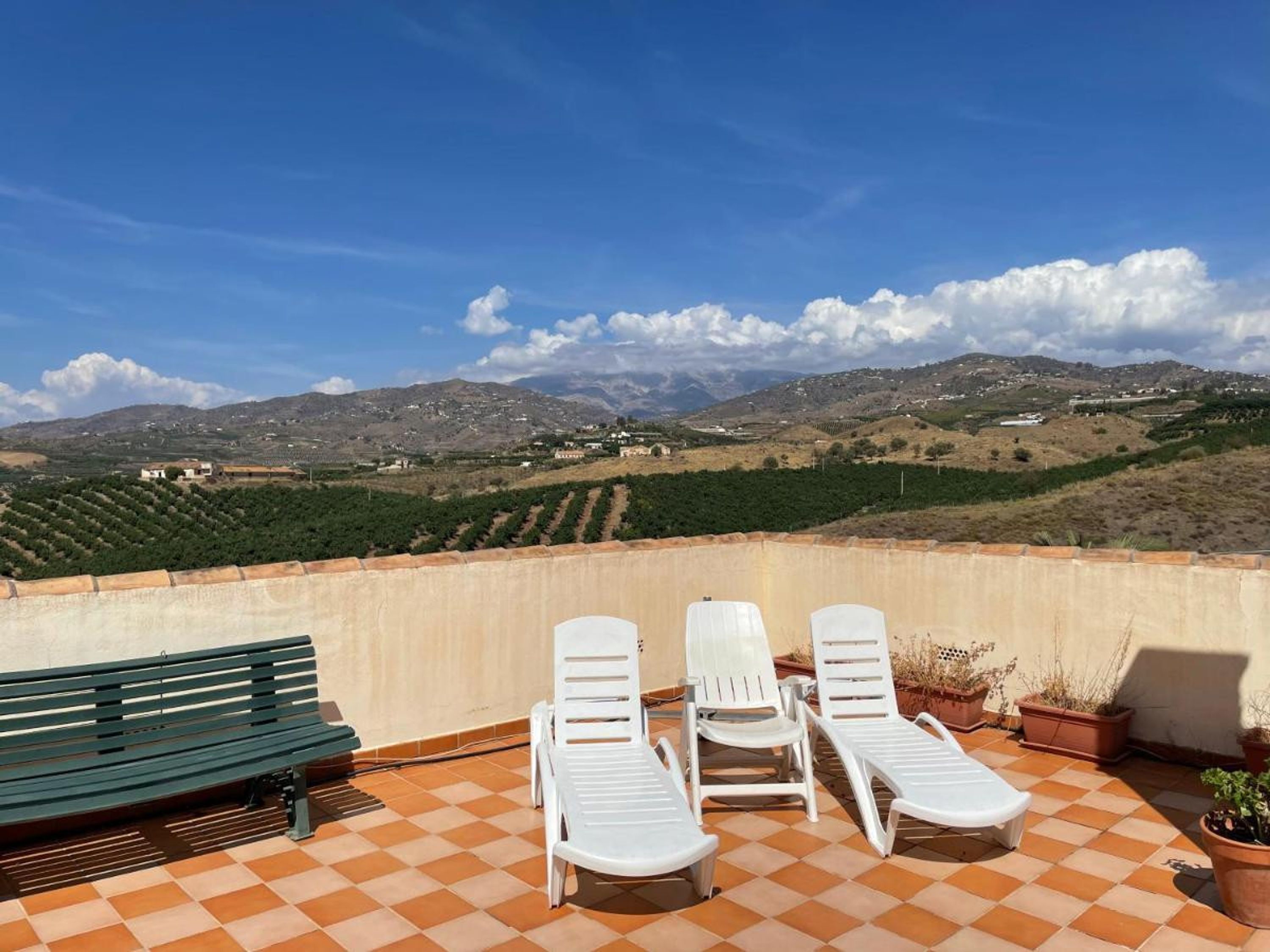 Eine Terrasse mit Schachbrettmuster, einer grünen Bank und weißen Liegestühlen mit Blick auf eine hügelige Landschaft.