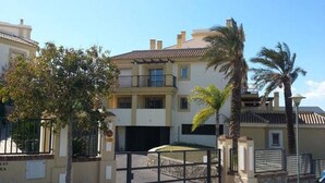 A multi-story residential building with a gated entrance, palm trees, and a clear blue sky. - Terrasol Tico Baviera Golf LAS Carmelitas (Velez-Malaga)