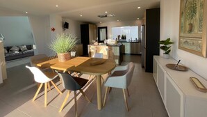 Moderni keittiö, jossa on keskeinen saareke, ruokailutila puupöydän ja -tuolien kera sekä olohuone, jossa on sohva ja kasvi.