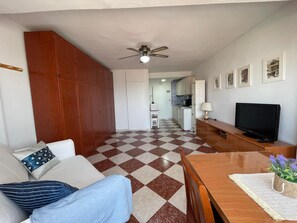A compact living space with a checkered floor, a wooden wardrobe, a sofa with cushions, a television, and a kitchenette in the background. - Terrasol Plazamar 4-9 (Velez-Malaga)