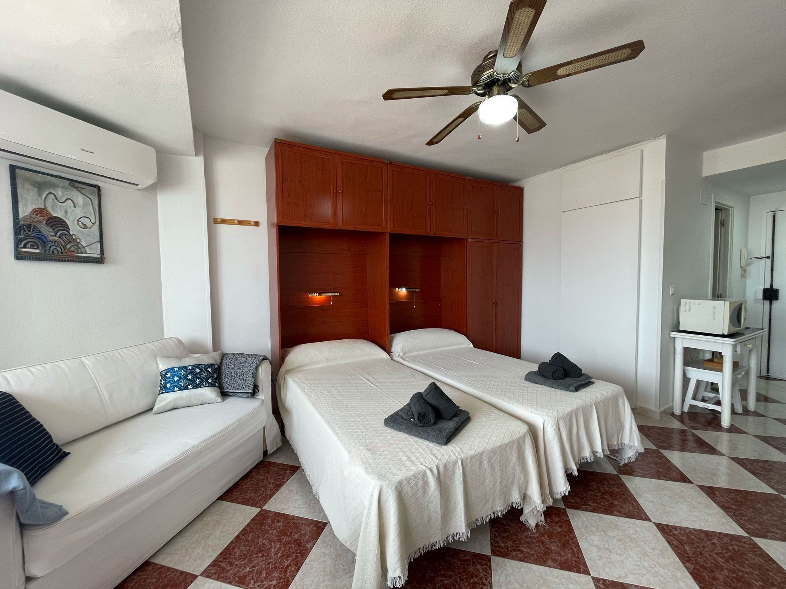 A hotel room with a white sofa, two single beds, a ceiling fan, a microwave, and a checkered floor.