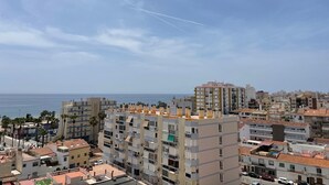 A coastal cityscape with multi-story buildings, a clear sky, and a visible contrail. - Terrasol Plazamar 4-9 (Velez-Malaga)