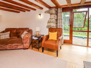 Una habitación con un sofá de cuero marrón, un sillón de cuero marrón, una pequeña mesa de madera con libros, una lámpara y una pared de piedra.