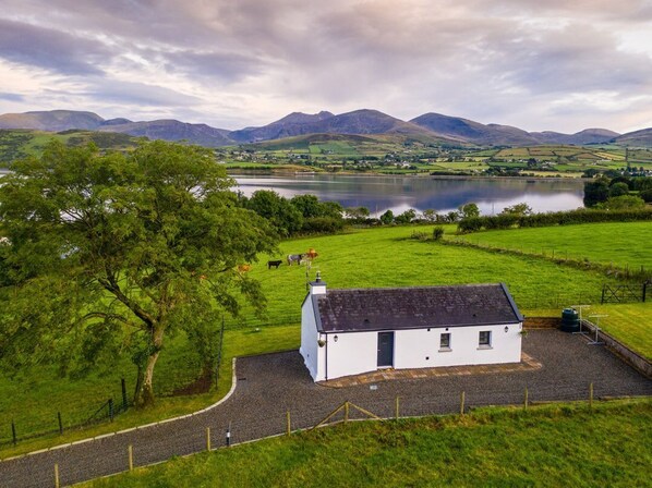 A white building with a black roof, surrounded by green fields and a lake, with mountains in the background.